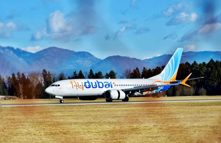 flydubai Boeing 737 Flugzeug auf Rollfeld vor malerischer Bergkulisse. Das weiße Flugzeug mit hellblauem und orangefarbenem Streifen trägt das 'flydubai' Logo. Im Vordergrund ist eine braune Grasfläche zu sehen, im Hintergrund bewaldete Berge unter blauem Himmel mit weißen Wolken. Das Bild illustriert einen Artikel über Gepäckrichtlinien bei flydubai, einschließlich Bestimmungen für Handgepäck, aufgegebenes Gepäck, Übergepäck, Sondergepäck, die Beförderung von Tieren und spezielle Regelungen für Kinder unter 2 Jahren, wie zusätzliches Freigepäck und die Mitnahme von Kinderwagen.