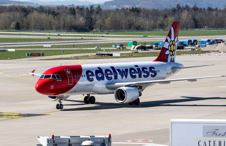 Auf dem Bild ist ein Flugzeug der Schweizer Fluggesellschaft Edelweiss Air auf einem Rollfeld zu sehen. Das Flugzeug hat eine markante rote Nase, und das Leitwerk ist mit dem bekannten Edelweiss-Blumen-Logo versehen. Im Hintergrund erstreckt sich ein Flughafenbereich mit grünen Wiesen und Bäumen sowie geparkten Fahrzeugen. Im Vordergrund steht ein Catering-Lkw, der das Flugzeug vermutlich für den bevorstehenden Flug vorbereitet. Diese Szene verweist auf das Thema des Blogartikels über die Edelweiss Handgepäck-Bestimmungen, in dem erklärt wird, welche Gegenstände im Handgepäck erlaubt sind, welche Masse und Gewichtsvorgaben einzuhalten sind und welche Regeln für Flüssigkeiten, elektronische Geräte oder Duty-Free-Artikel gelten. Wer mit Edelweiss reist, sollte gut vorbereitet sein, um eine reibungslose Abfertigung und stressfreie Flugerfahrung zu haben.