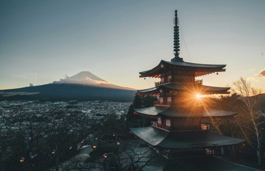 Japan Reise: Die berühmte Pagode von Chureito mit dem majestätischen Mount Fuji im Hintergrund bei Sonnenaufgang. Das Bild zeigt die ikonische fünfstöckige Pagode in Arakurayama Sengen Park, Fujiyoshida, deren rote Holzstrukturen kontrastreich vor dem Hintergrund des schneebedeckten Fuji erstrahlen. Die aufgehende Sonne beleuchtet die Pagode von hinten, wodurch die Silhouetten der Dachebenen hervorgehoben werden und ein warmes, goldenes Licht die Umgebung erfüllt. Unten erstreckt sich die Stadt Fujiyoshida, die in den sanften Hügeln eingebettet ist. Dieses Bild fängt die Essenz einer Japan Reise ein, mit seiner einzigartigen Verbindung von traditioneller Architektur und atemberaubender Natur