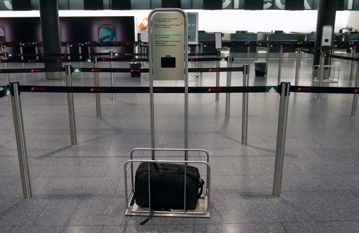 Das Bild zeigt eine leere Warteschlange am Check-in-Schalter von SWISS Airlines. Im Vordergrund steht eine Handgepäck-Schablone aus Metall, die dazu dient, die Grösse des Handgepäcks zu überprüfen. Ein schwarzer Rucksack befindet sich innerhalb des Rahmens, der die maximal erlaubte Grösse von 55 x 40 x 23 cm für SWISS Handgepäck darstellt. Der Rahmen ist auf einem Metallständer befestigt, über dem ein Schild angebracht ist. Auf dem Schild steht in mehreren Sprachen, dass Handgepäck, das nicht in den Rahmen passt, aufgegeben werden muss. Im Hintergrund sind weitere Warteschlangenabsperrungen zu sehen, die ebenfalls mit „SWISS“-Logos versehen sind. Der Check-in-Bereich im Hintergrund ist leer, was auf eine ruhige Zeit am Flughafen hindeutet.