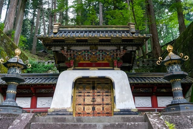 Der prachtvolle Eingang zum Toshogu-Schrein in Nikkō, einer der bekanntesten und kunstvollsten Tempelanlagen Japans. Die massive Holztür ist mit goldenen Ornamenten verziert und zeigt detaillierte Schnitzereien, die den Reichtum und die Bedeutung des Schreins betonen. Links und rechts vom Eingang stehen verzierte Laternen aus Metall, die dem Schrein eine majestätische und historische Atmosphäre verleihen. Der Schrein selbst ist von einem dichten, grünen Wald umgeben, was ihm eine friedvolle und spirituelle Aura verleiht. Der Toshogu-Schrein ist die Grabstätte von Tokugawa Ieyasu, dem Gründer des Tokugawa-Shogunats, und zählt zu den wichtigsten Sehenswürdigkeiten für eine Japan Rundreise.