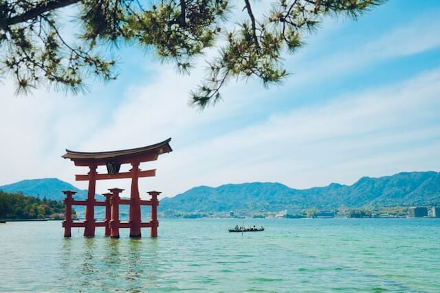 Japan Reise: Das berühmte rote Torii-Tor des Itsukushima-Schreins auf der Insel Miyajima, das scheinbar auf dem Wasser schwebt. Das große, leuchtend rote Tor ragt aus dem türkisgrünen Wasser des Meeres empor und bildet einen beeindruckenden Kontrast zur natürlichen Umgebung. Im Hintergrund erstrecken sich bewaldete Hügel und sanfte Bergketten, die die ruhige Atmosphäre des Ortes verstärken. Das Tor ist teilweise von den überhängenden Ästen eines Pinienbaums eingerahmt, der das Bild von oben säumt und dem Anblick eine zusätzliche Tiefe verleiht. Auf dem Wasser sieht man ein kleines traditionelles Ruderboot, das auf das Torii-Tor zusteuert, was die spirituelle Bedeutung und die Anziehungskraft dieses Ortes für Besucher unterstreicht. Dieses Bild fängt die Magie und den Frieden von Miyajima ein, einer der bekanntesten Sehenswürdigkeiten Japans und ein Highlight jeder Japan Reise, zusammen mit Hiroshima, dem Ort des Gedenkens an die Vergangenheit