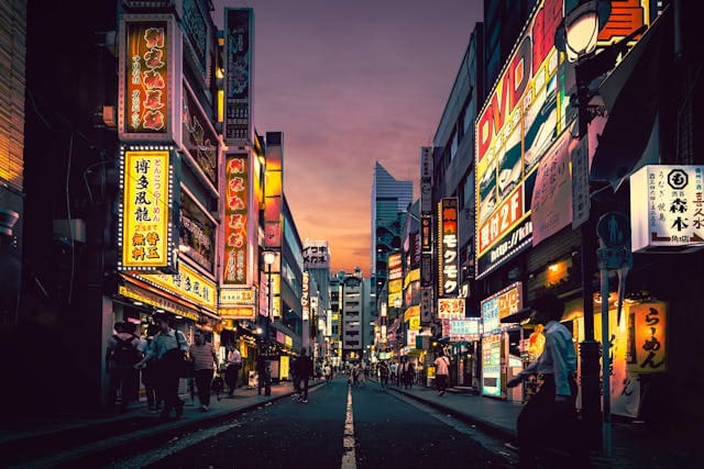 Eine belebte, neonbeleuchtete Straße in Tokio bei Sonnenuntergang, die das pulsierende Nachtleben und die lebendige Atmosphäre der Stadt einfängt. Auf beiden Seiten der schmalen Straße sind hohe Gebäude mit leuchtenden Reklametafeln und bunten Schildern zu sehen, die in Kanji und lateinischen Buchstaben geschrieben sind und verschiedene Geschäfte, Restaurants und Bars anpreisen. Menschen schlendern entlang der Gehwege, tauchen ein in das bunte Lichtermeer und die dynamische Szenerie. Das Bild vermittelt den einzigartigen Mix aus traditioneller japanischer Architektur und modernen, urbanen Einflüssen, der Tokio zu einem beliebten Startpunkt für eine Japan Rundreise macht. Dieses Viertel ist typisch für die energiegeladene Metropole, in der Tradition und Moderne nebeneinander existieren.