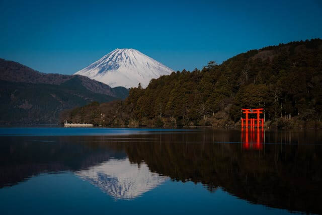 Japan Reise: Der beeindruckende Anblick des schneebedeckten Mount Fuji, der sich majestätisch im Hintergrund erhebt, mit ruhigem, tiefblauem Himmel darüber und klarer Sicht auf die ikonische Silhouette des höchsten Berges Japans. Im Vordergrund liegt der Ashi-See, dessen ruhige Wasseroberfläche das Bild des Fuji perfekt spiegelt und eine friedliche, symmetrische Komposition schafft. Auf der rechten Seite des Sees ragt das leuchtend rote Torii-Tor des Hakone-Schreins aus dem Wasser empor und bildet einen starken Kontrast zur umliegenden Natur. Dichte grüne Wälder bedecken die Ufer des Sees und rahmen das Bild ein, während die Berge im Hintergrund die Szenerie abrunden. Dieses Bild symbolisiert die Ruhe und natürliche Schönheit von Hakone, einem beliebten Ziel auf Japan Reisen, bekannt für seine heissen Quellen (Onsen), traditionelle Ryokan-Unterkünfte und spektakuläre Fuji-Ausblicke.