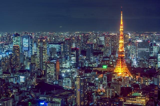 Japan Reise: Nächtliche Skyline von Tokio, aufgenommen aus der Vogelperspektive. Im Zentrum des Bildes steht der ikonische Tokyo Tower, ein hoher, funkelnder Turm in leuchtendem Orange, der bei Dunkelheit die Stadt erhellt und an den Pariser Eiffelturm erinnert. Der Turm ist in kräftigem, warmem Licht beleuchtet und hebt sich majestätisch von der kühlen, dunkelblauen Nachtkulisse ab. Rund um den Tokyo Tower erstreckt sich das dichte Stadtgebiet Tokios, eine scheinbar endlose Landschaft aus hohen, modernen Wolkenkratzern, Bürogebäuden und Wohnhäusern, die ebenfalls von tausenden kleinen weißen, blauen und gelben Lichtern glitzern. Die Fenster und Lichter der Gebäude reflektieren die Lebendigkeit der Stadt, während die Straßen darunter in einem Netz aus Lichtern schimmern und sich durch das Bild ziehen. Die gesamte Szene vermittelt die Energie und Dynamik der japanischen Hauptstadt, die nie zu schlafen scheint. Dieses Bild fängt die Essenz der Tokio-Etappe einer Japan Reise ein – von den vibrierenden, hell erleuchteten Straßen der Metropole bis hin zu den vielen Sehenswürdigkeiten, die Tokio zu bieten hat, wie der Tokyo Skytree, die geschäftige Shibuya-Kreuzung, der historische Senso-ji Tempel in Asakusa und die Oase der Ruhe im Shinjuku Gyoen Nationalpark. Ein lebendiges, modernes Tokio, das dennoch voller Geschichte und Kultur steckt