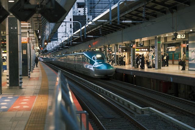 Ein glänzend türkiser Shinkansen-Hochgeschwindigkeitszug steht auf einem Bahnsteig einer modernen und gut beleuchteten Bahnstation in Japan, bereit zur Abfahrt. Der Zug hat eine aerodynamische Form mit einer langen, abgerundeten Nase und großen Fenstern, was ihm ein futuristisches Aussehen verleiht. Auf der linken Seite des Bahnsteigs ist ein gelbes Sicherheitsband zu sehen, das den Abstand zur Bahnlinie markiert. Verschiedene Schilder und Anzeigetafeln über den Gleisen zeigen Nummern und Informationen für Reisende an, während einige Fahrgäste auf den Bahnsteigen warten. Die Bahnstation ist sauber und modern, mit breiten Gängen und Überdachungen aus Metall, die die gesamte Länge des Bahnsteigs abdecken. Im Hintergrund sind hohe Gebäude zu erkennen, die den urbanen Charakter des Ortes betonen. Dieses Bild vermittelt die Effizienz und den Komfort des Zugverkehrs in Japan, ideal für eine Japan Rundreise mit dem Japan Rail Pass