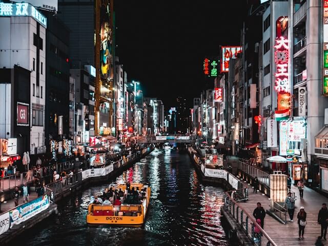 Japan Reise: Nachtansicht des belebten Dotonbori-Viertels in Osaka mit dem beleuchteten Kanal im Zentrum. Auf dem Wasser fährt ein gelbes Ausflugsboot mit Touristen, die die farbenfrohen Neonlichter und Leuchtreklamen bestaunen, die die Straße säumen. Links und rechts des Kanals erstrecken sich dicht gedrängte Gebäude, deren Fassaden mit auffälligen, bunten Schildern und Reklamen in Kanji-Zeichen und grellen Farben geschmückt sind. Die Glico-Man-Leuchtreklame und zahlreiche weitere riesige Werbetafeln tragen zur lebendigen, energiegeladenen Atmosphäre des Viertels bei. Entlang der Uferpromenade schlendern Fußgänger, die die lokale Straßenküche genießen und die einzigartige Stimmung von Dotonbori auf sich wirken lassen. Dieses Bild zeigt die Essenz von Osaka, dem kulinarischen Herzen Japans, das bekannt ist für seine Spezialitäten wie Takoyaki und Okonomiyaki, und bietet Besuchern ein intensives urbanes Erlebnis voller Lichter und Leben