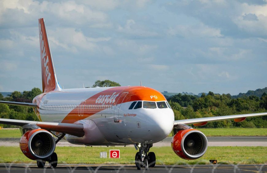 Ein easyJet-Flugzeug, das auf einem Rollfeld steht und sich auf den Start oder die Landung vorbereitet. Das Flugzeug ist in den typischen Farben von easyJet gestaltet, mit einer weißen Grundfarbe und kräftigen orangefarbenen Akzenten, die das Logo und die Triebwerke zieren. Im Vordergrund sieht man den linken Motor mit seinem rotierenden Turbinenrad, während sich das Hauptfahrwerk und die Nase des Flugzeugs in Richtung der Startbahn erstrecken. Im Hintergrund ist eine grüne Landschaft mit Bäumen sowie ein leicht bewölkter Himmel zu sehen. Dieses Bild veranschaulicht den Blogartikel über die easyJet Handgepäck-Bestimmungen und bietet einen visuellen Bezug zur Fluggesellschaft, die Reisende dazu anhält, sich gut auf die Handgepäckvorschriften vorzubereiten, um eine reibungslose Reise zu gewährleisten