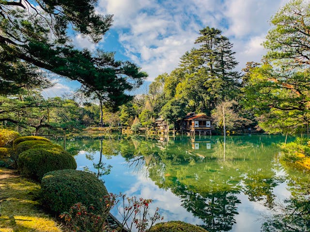 Kenroku-en Garten in Kanazawa, einer der berühmtesten Landschaftsgärten Japans, an einem klaren Tag mit ruhigem, spiegelndem Wasser, das die umgebende Vegetation reflektiert. Im Vordergrund sorgfältig gepflegte Sträucher, im Hintergrund hohe Bäume und traditionelle japanische Gebäude, die das historische und friedliche Ambiente dieses Gartens betonen. Dieser Garten zeigt die perfekte Harmonie der japanischen Gartenkunst und ist ein beliebter Ort für Besucher auf einer Japan Rundreise, die Entspannung und Natur genießen möchten.