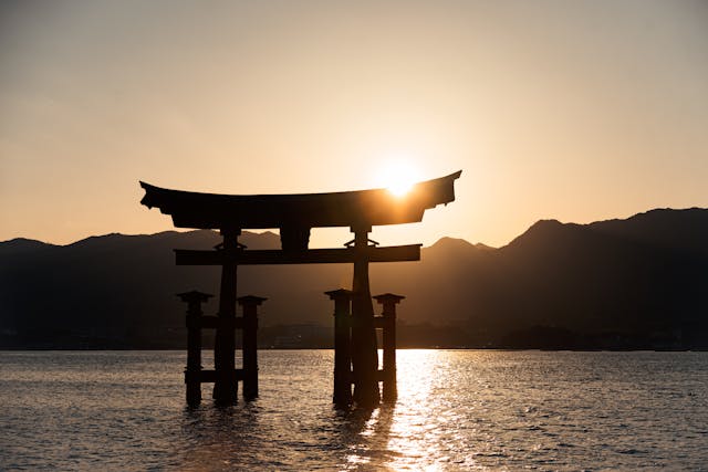 as berühmte Torii-Tor des Itsukushima-Schreins auf der Insel Miyajima, Japan, im Sonnenuntergang. Das Tor scheint über dem Wasser zu schweben, da es bei Flut teilweise im Meer steht. Die untergehende Sonne strahlt golden durch die Torbalken und wirft ein sanftes Licht auf die Wasseroberfläche, während die Silhouetten der umliegenden Berge die friedvolle und spirituelle Atmosphäre der Szenerie verstärken. Miyajima, auch als „Insel der Götter“ bekannt, ist ein spiritueller und fotogener Ort, der Besucher auf einer Japan Rundreise in den Bann zieht