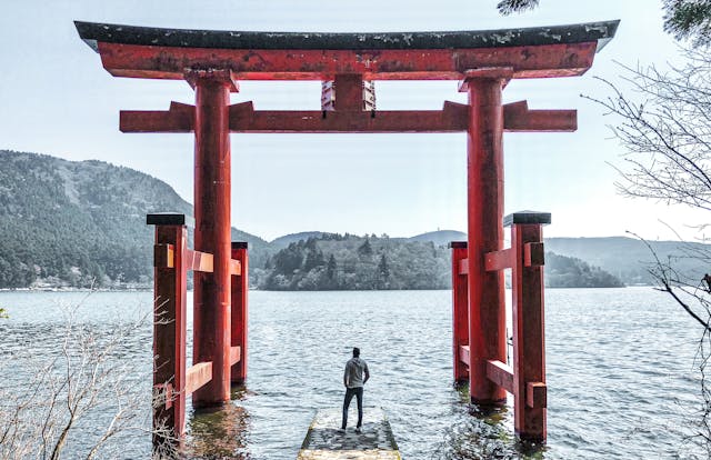 Ein Reisender steht vor dem ikonischen roten Torii-Tor des Hakone-Schreins, das direkt im Wasser des Ashi-Sees (Ashinoko) verankert ist. Das große Tor markiert den Übergang zu einer heiligen Stätte und bietet eine malerische Kulisse, die die japanische Kultur und Spiritualität widerspiegelt. Im Hintergrund ist die bewaldete Landschaft und die sanfte Hügellandschaft rund um den See zu sehen, die eine friedliche und beeindruckende Atmosphäre schaffen. Diese Szene symbolisiert die Verbindung von Mensch, Natur und Tradition und ist ein beliebtes Fotomotiv für Reisende auf ihrer Japan Rundreise