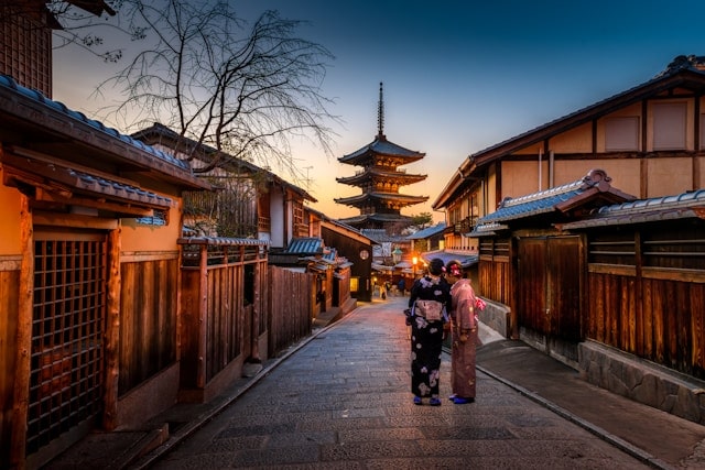 Eine abendliche Straßenszene im historischen Stadtteil Higashiyama in Kyoto, Japan, zeigt zwei Menschen in traditionellen Kimonos, die entlang einer ruhigen gepflasterten Straße schlendern. Im Hintergrund erhebt sich die markante Pagode des Yasaka-Schreins, die in warmem Licht erstrahlt und die traditionelle japanische Architektur der Umgebung ergänzt. Die schmalen Gassen und die hölzernen Machiya-Häuser verstärken das authentische Ambiente dieser alten Kaiserstadt, die reich an kulturellen und spirituellen Erlebnissen ist – ein Muss auf jeder Japan Rundreise.